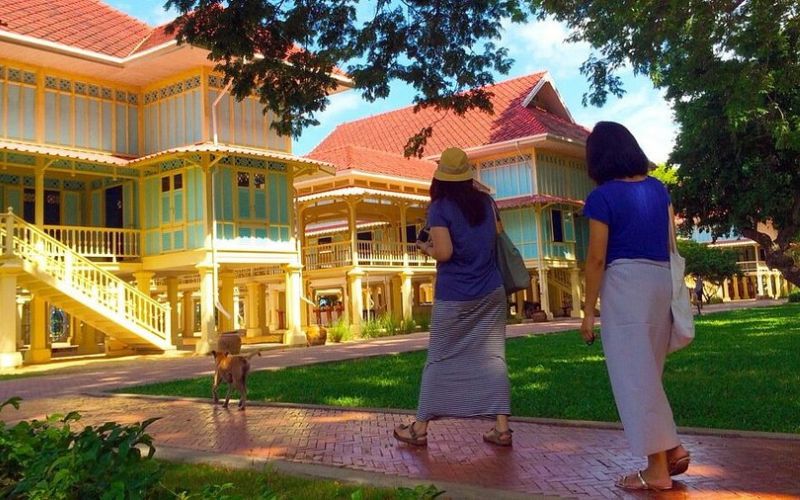 Two women walking through the garden of Klai Kangwon Palace, a traditional Thai royal residence with yellow wooden buildings and red roofs
