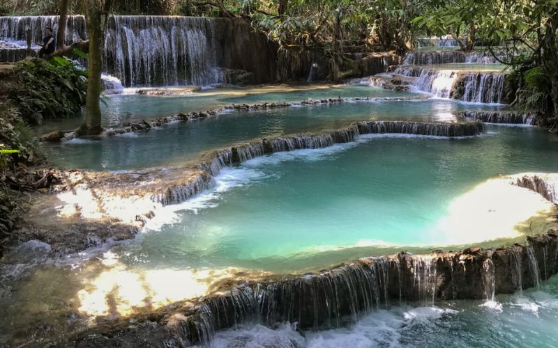 Turquoise-blue pools of water cascading down limestone tiers at Kuang Si Waterfalls in Luang Prabang, Laos, surrounded by lush greenery.