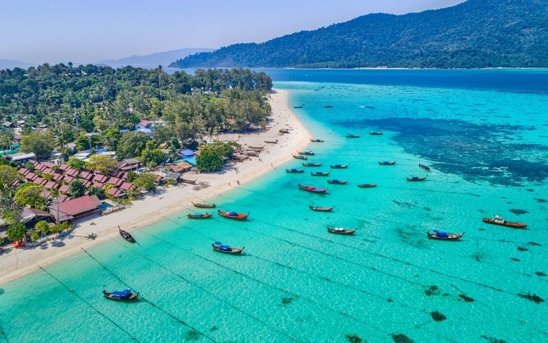 An aerial view of a pristine white-sand beach and resort area, with numerous traditional longtail boats floating on incredibly clear, bright turquoise shallow water that stretches towards a lush, green mountainous island backdrop.