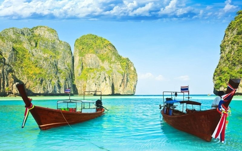 Two traditional Thai long-tail boats floating on crystal-clear turquoise water surrounded by towering limestone cliffs and lush greenery at Koh Lanta, Thailand.