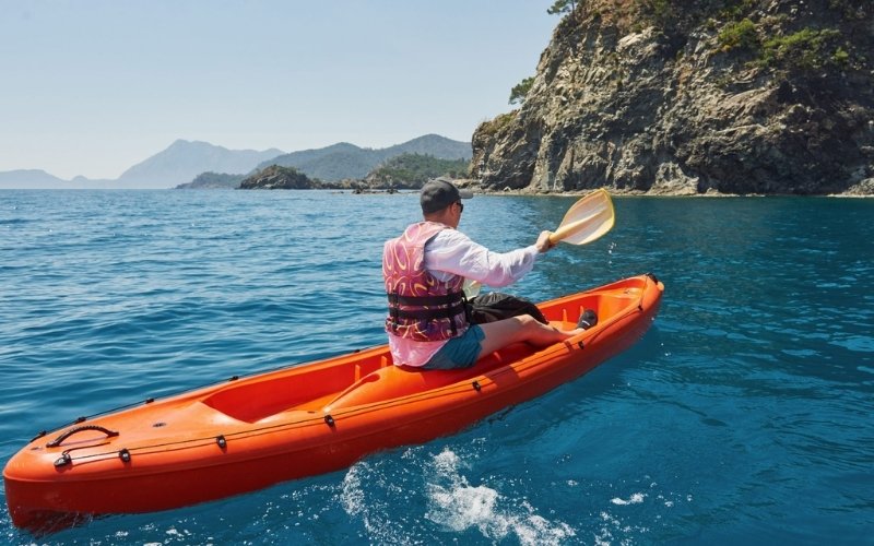 A person kayaking in a bright orange kayak on clear blue water near rocky cliffs and green hills at Phi Phi Island, Thailand