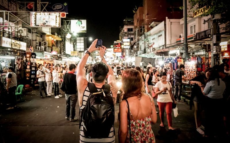 A night view of Khao San Road in Bangkok from behind a couple, with a man in a striped tank top holding up his phone to take a photo of the brightly lit street filled with market stalls, neon signs, and crowds of people.