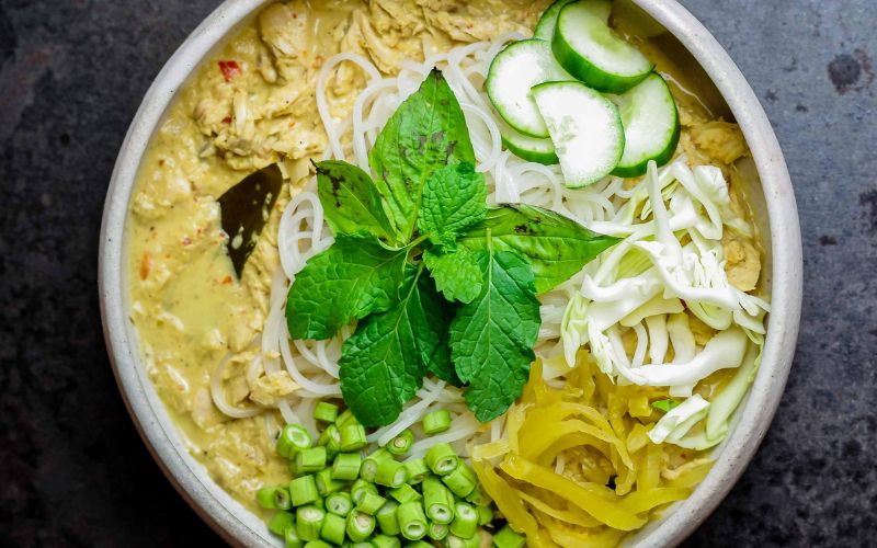 A bowl of khanom jeen nam ya, rice noodles topped with fish curry, mint, cucumber, green beans, and pickled mustard greens.