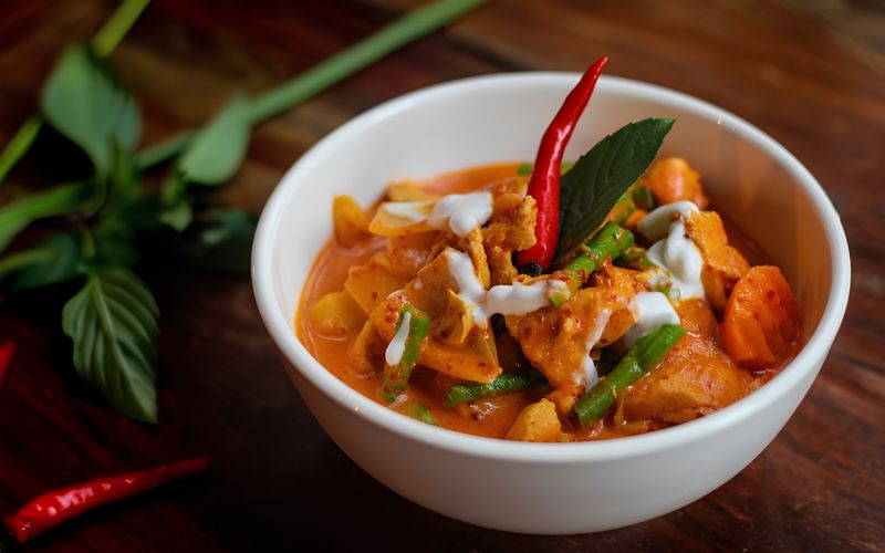 A bowl of Khmer red curry with chicken, carrots, green beans, and coconut milk, garnished with basil and red chili.