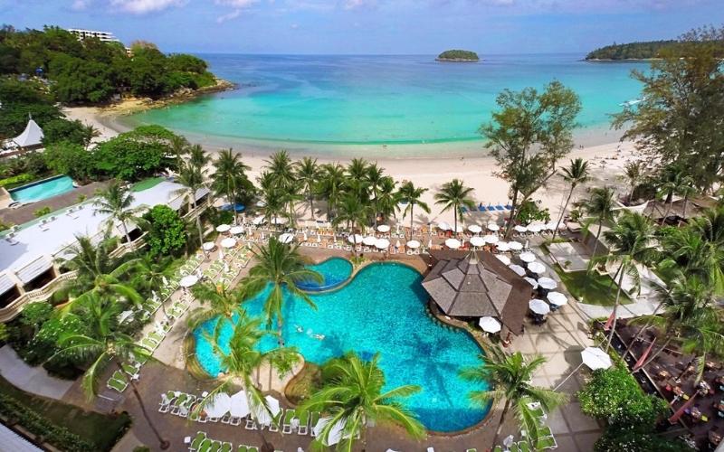 A high-angle view overlooking a resort swimming pool complex and the stunning Kata Beach in Phuket, Thailand. The resort features a large, winding turquoise pool surrounded by sun loungers, palm trees, and a central tiki-style bar. Beyond the resort, the white-sand beach meets the crystal-clear, azure waters of the Andaman Sea, with a small island visible on the horizon.