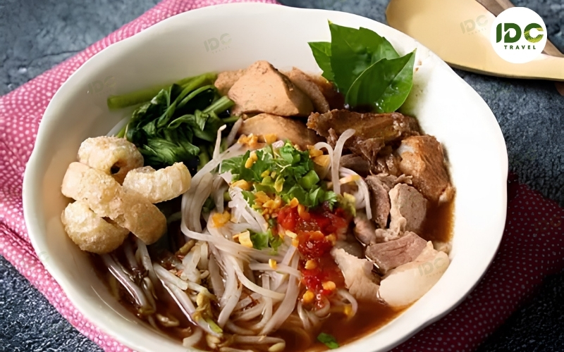 A bowl of kuai tiao ruea, Thai boat noodles with rice noodles, pork, herbs, bean sprouts, and pork rinds in a dark broth.