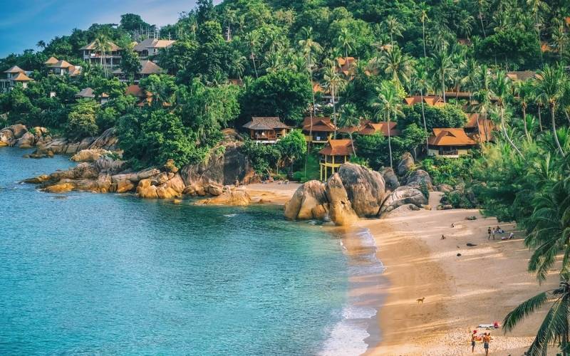 Aerial view of a tropical beach in Koh Samui, Thailand, with golden sand, turquoise water, and hillside resorts surrounded by lush palm trees.