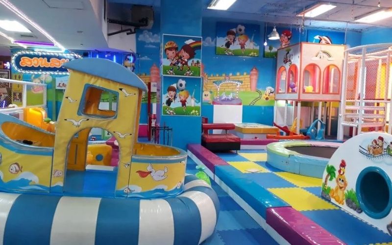 A brightly decorated indoor soft play area or kids' café, featuring colorful mats, a boat-shaped structure, a trampoline, and tunnels, designed for family entertainment in Bangkok or Chiang Mai.