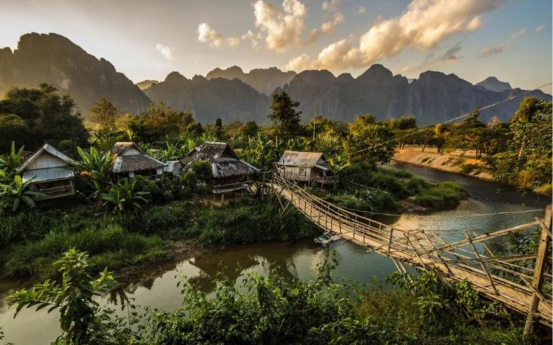 A peaceful rural village in Laos with wooden huts surrounded by lush greenery, a small wooden bridge crossing a river, and dramatic mountains in the background under a golden sunset sky.