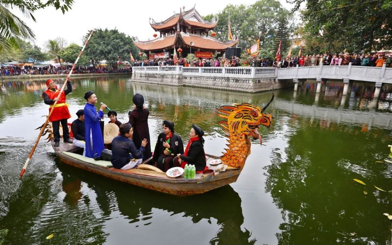 Traditional Quan Ho singers performing on a dragon-headed boat in a lake during the Lim Festival, with a tiered pagoda and crowds in the background.