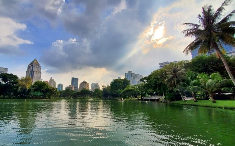 A wide, scenic view of the central lake in Lumpini Park, Bangkok, showcasing lush greenery and palm trees contrasting with the modern city skyline under a dramatic, cloudy sky.