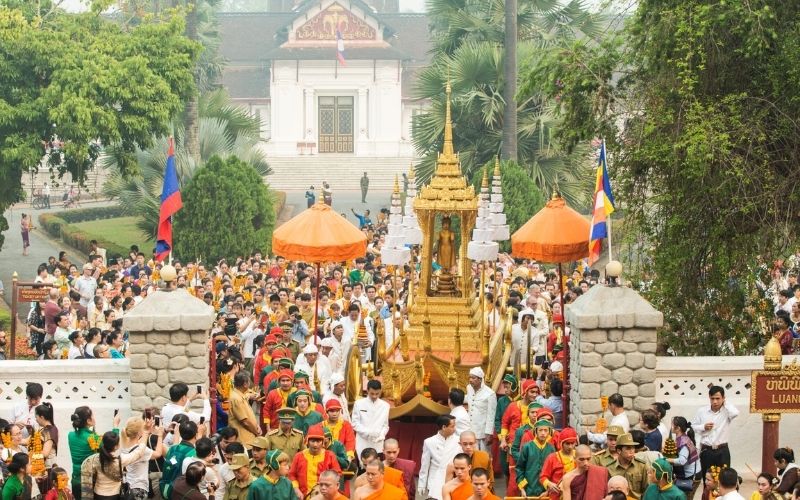 Crowds celebrating Lao New Year Festival in Luang Prabang, with traditional golden procession floats, people dressed in colorful attire, and a temple in the background.