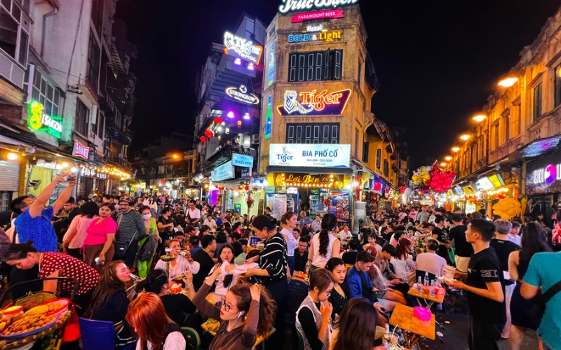 Locals and travelers enjoy cold Hanoi Beer on plastic stools along the buzzing sidewalks of Ta Hien Street, with street lights, laughter, and glasses clinking over freshly poured beer.