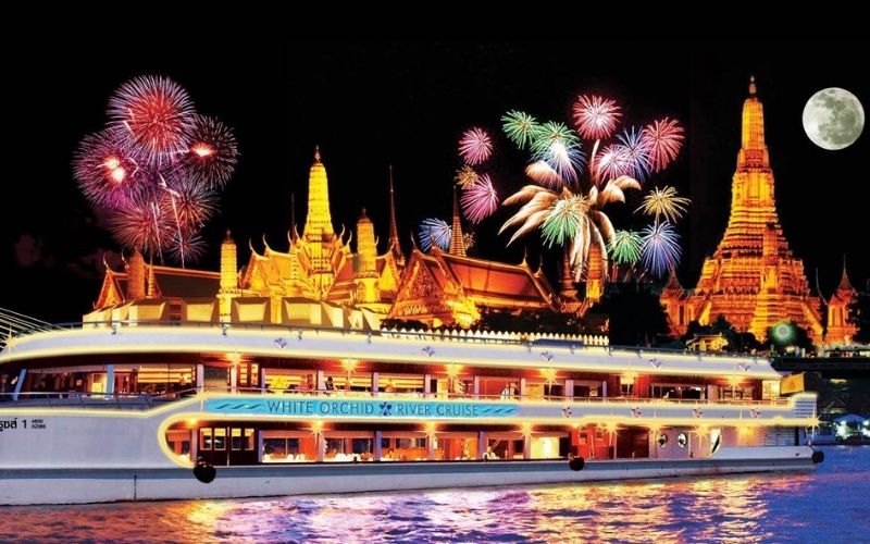 A brightly lit river cruise ship sails past illuminated Thai temples at night with colorful fireworks bursting in the sky and a full moon shining above.