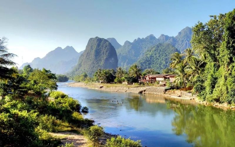 A serene river winding through lush greenery with tropical trees, traditional houses, and dramatic limestone mountains under a clear blue sky.
