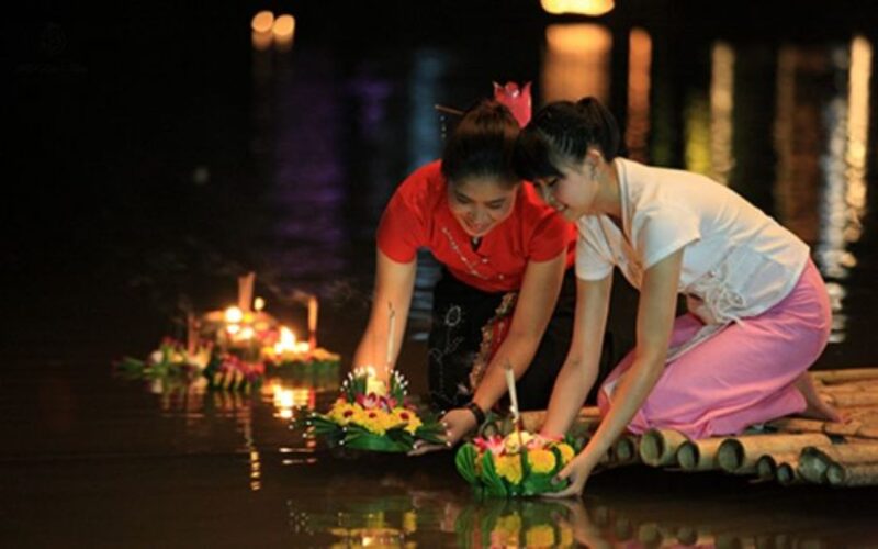 Two women dressed in traditional Thai attire gently release their handmade Krathongs onto the river during the Loy Krathong Festival
