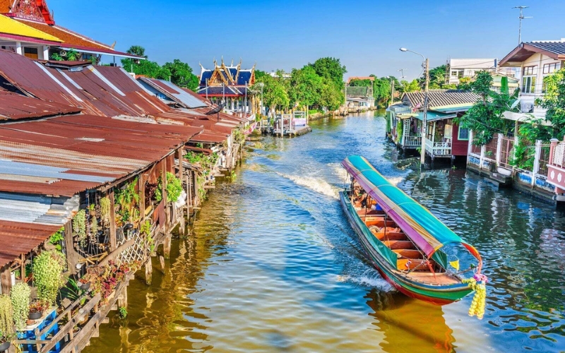 A long-tail boat moving through a peaceful canal surrounded by traditional wooden homes in old Bangkok.