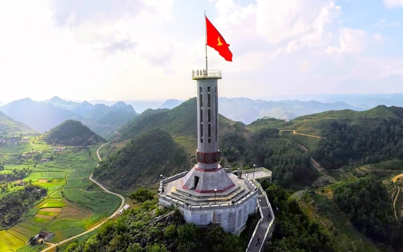 Lung Cu Flag Tower standing proudly on a hilltop in Ha Giang, Vietnam, with a large red national flag waving against a clear blue sky and panoramic mountain views in the background.
