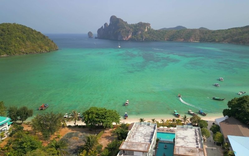 Aerial view of Loh Dalum Bay on Phi Phi Island, Thailand, showing turquoise water, white sand beach, boats on the bay, and lush green hills in the distance.