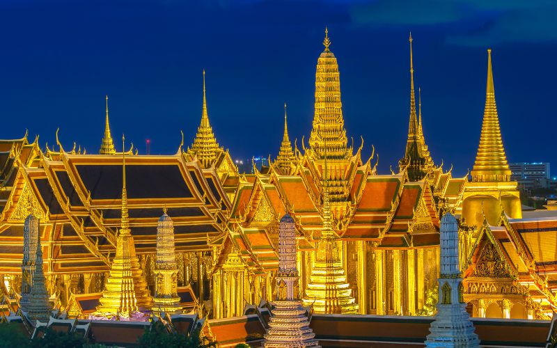 A sweeping nighttime view of the Grand Palace complex in Bangkok, where dozens of multi-tiered golden chedis and ornate temple roofs glow with bright warm lights against a deep blue evening sky.