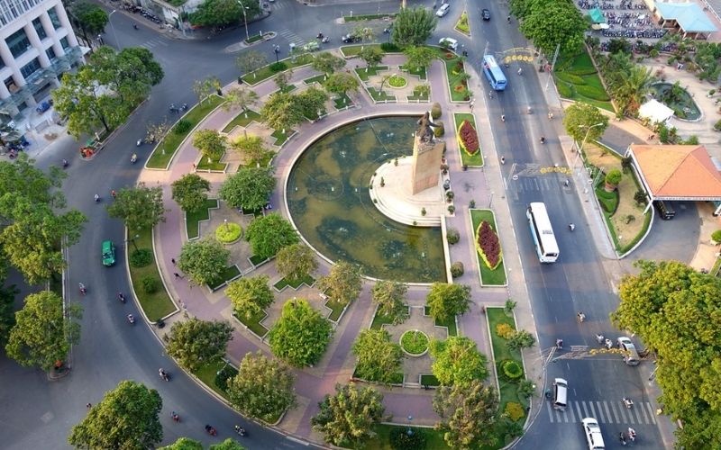 An elevated daytime view of Me Linh Square in Ho Chi Minh City, showing the circular green plaza with the Tran Hung Dao statue, surrounded by busy roads and lush trees leading toward the river.