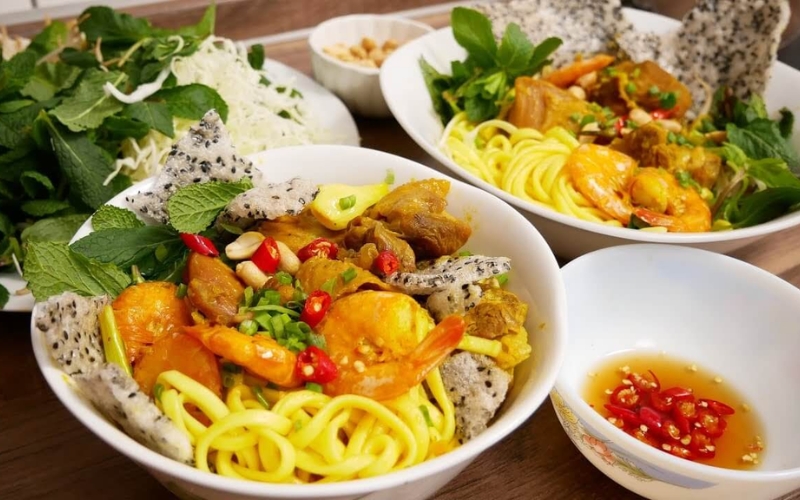 A bowl of Mi Quang noodles topped with shrimp, pork, quail egg, herbs, and crispy rice cracker, served in Da Nang