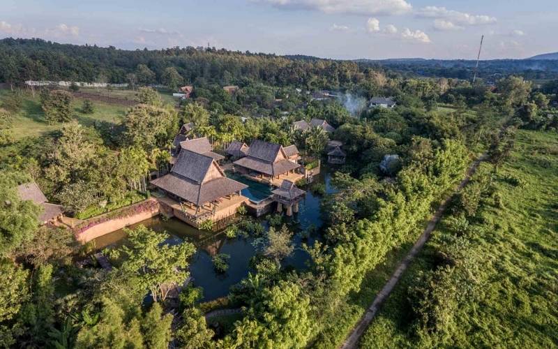 A high-angle aerial shot of a beautifully designed resort or café complex in Mae Rim, Chiang Mai, Thailand, featuring traditional Lanna-style buildings with dark, multi-tiered thatched roofs built over or surrounded by a large pond. The complex is nestled within a dense, lush green landscape of trees and fields, highlighting its secluded, natural setting.
