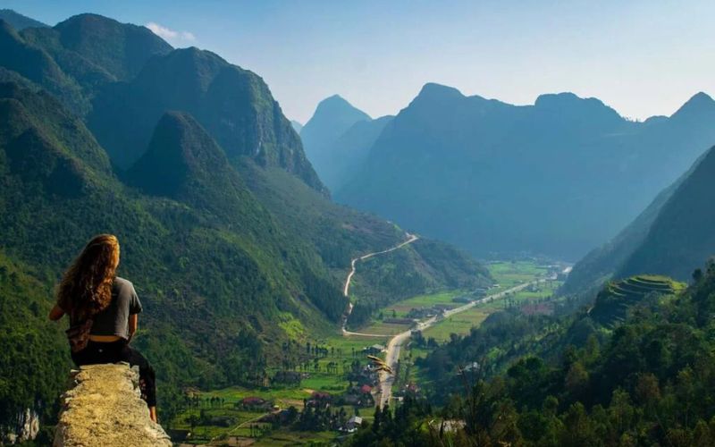 Female traveler sitting on a rock ledge overlooking a stunning valley surrounded by dramatic limestone mountains along the Ha Giang Loop in northern Vietnam