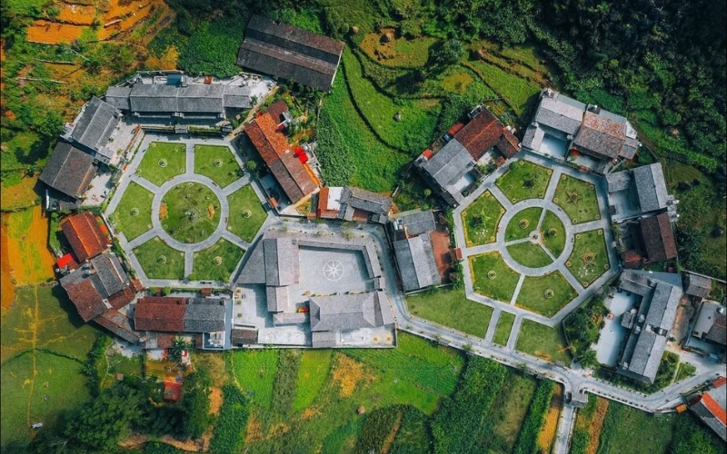 A panoramic view of Meo Vac Village, nestled in a deep valley surrounded by rugged limestone mountains, with traditional stilt houses, winding roads, and terraced fields creating a picturesque highland scene.
