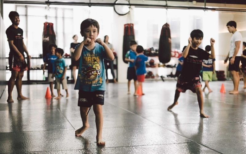 Several young children participate in a beginner's Muay Thai class, practicing fighting stances barefoot on a gym floor with instructors and punching bags visible in the background.