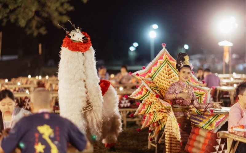 A festive night scene at a New Year celebration or cultural event in Chiang Mai, Thailand. In the foreground, a performer in an elaborate, colorful Lanna-style costume with large, winged back pieces smiles at the camera, standing next to a performer wearing a tall, shaggy white costume resembling a mythical creature or dragon. People are seated at low bamboo tables in the background.