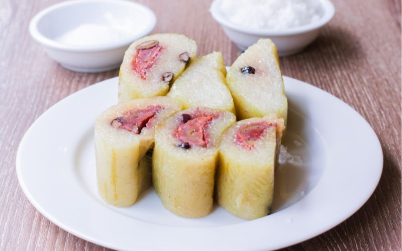 Sliced nom ansom chek, sticky rice and banana cake wrapped in banana leaf, served on a white plate with coconut.