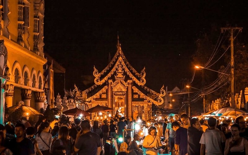 A vibrant night scene of a crowded walking street or night market in Chiang Mai, Thailand. In the background, a beautifully illuminated traditional Lanna-style temple gate or building entrance (likely Wat Chedi Luang or Wat Phra Singh area) glows gold against the dark sky. The foreground is filled with people browsing stalls under warm streetlights.