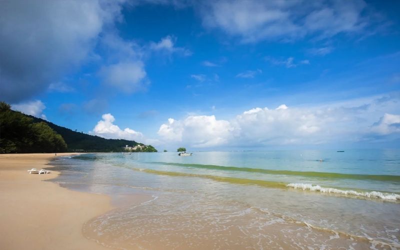A wide, bright photograph of Nai Yang Beach, featuring wide, light-colored sand, clear turquoise water with gentle waves, and a steep, dark green forested hillside meeting the sea under a bright blue sky with large white cumulus clouds.