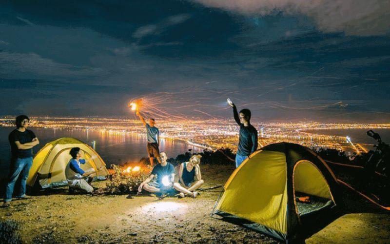 Travelers camping and enjoying a night picnic on Hai Van Pass, overlooking Da Nang city lights under a starry sky