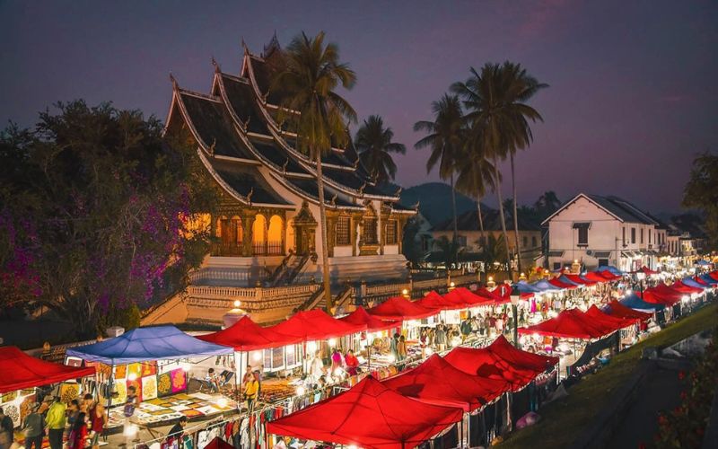 Vibrant night market in Luang Prabang, Laos, with colorful stalls selling textiles, crafts, and street food, illuminated by soft lighting against a dusky sky.