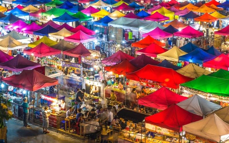 A high-angle view looking down on a bustling night market filled with a colorful patchwork of illuminated red, blue, pink, and yellow tent roofs.