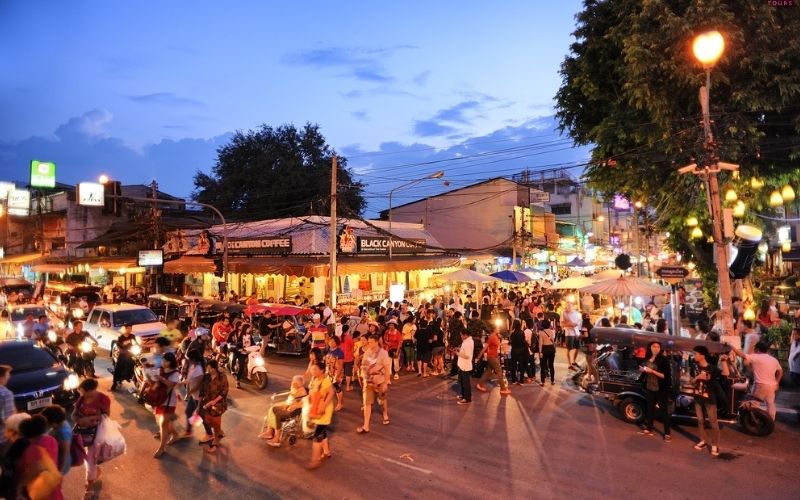 A vibrant and crowded street scene at dusk in Chiang Mai, Thailand, likely a night bazaar or walking street. The street is bustling with pedestrians, motorbikes, and tuk-tuks. Numerous market stalls with illuminated awnings line both sides of the road, and storefronts like "Black Canyon Coffee" are visible. Warm streetlights and hanging decorative lights add to the lively atmosphere.