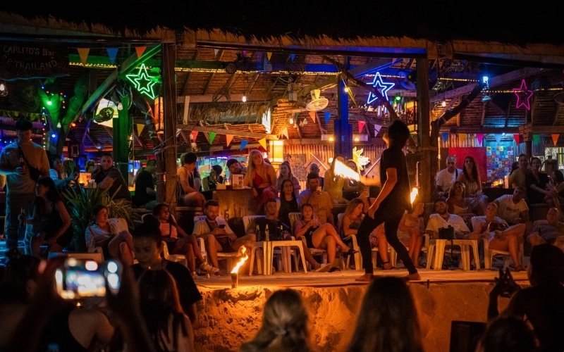 A lively beach bar scene at night on Phi Phi Island, with people watching a fire dancing performance under colorful neon lights and festive decorations.