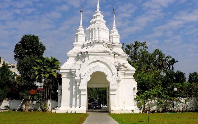 A striking white temple gate with traditional Thai Lanna architectural features, including pointed spires, set against a blue sky with white clouds. A paved walkway leads through the arched entrance, which is flanked by manicured green lawns and lush tropical trees, likely leading to Wat Suan Dok or a similar temple near the Nimman area of Chiang Mai.
