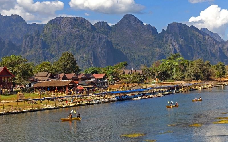 A scenic riverside view with kayakers paddling on the Nam Song River, traditional wooden houses along the shore, and majestic limestone mountains in the background.