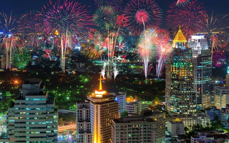 A spectacular display of colorful fireworks exploding over the illuminated skyscrapers of a city skyline in Thailand during New Year celebrations.