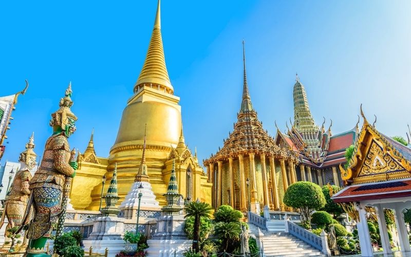 A bright, sunny photograph of the Grand Palace and Wat Phra Kaew (Temple of the Emerald Buddha) in Bangkok, Thailand. The scene is dominated by brilliant gold structures, including a large, conical golden stupa and ornate royal halls with tiered roofs, set against a clear blue sky. A massive, colorful guardian figure (Yaksha) stands prominently on the left side of the frame.