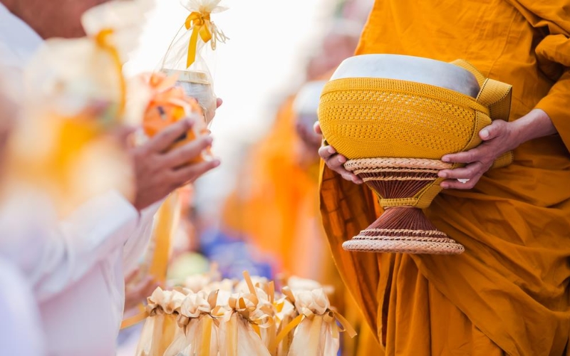 A Buddhist monk holds a traditional alms bowl as local devotees, dressed in white, offer carefully prepared food and gifts wrapped in saffron ribbons