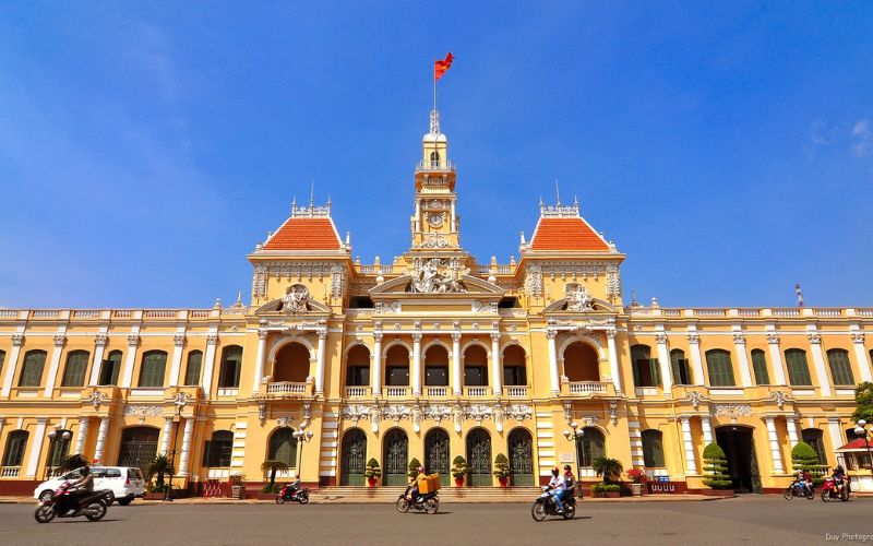 A view of Nguyen Hue Walking Street in Ho Chi Minh City at dusk, featuring the prominent bronze statue of President Ho Chi Minh in the left foreground. Behind the statue, the historic French colonial-style People's Committee Building (City Hall) is illuminated, with the Vietnamese flag flying from its central tower. A large, modern office building is visible on the right, contrasting with the historic structures.