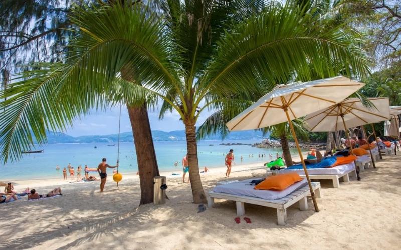 Beach scene with palm trees, sun loungers, and umbrellas along a sandy shore, with people relaxing and swimming in clear blue ocean water.