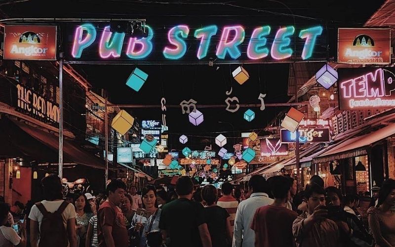 A nighttime photo of the bustling Pub Street in Siem Reap, Cambodia, illuminated by a large, colorful neon sign and numerous bar signs, with a dense crowd of people walking down the street.