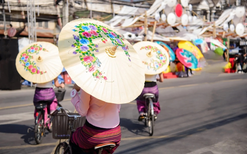 A close-up, back-view photo of three women in pink and purple traditional Thai outfits riding bicycles in a parade at the Bo Sang and Sankhampaeng festival. They are holding large, white parasols beautifully hand-painted with green, pink, and purple flowers.