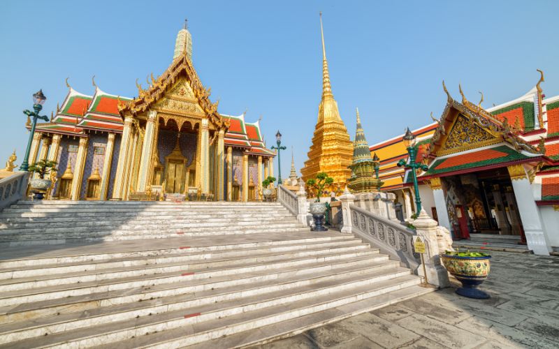 A view from the base of a wide set of light marble steps leading up toward the intricately detailed Phra Mondop (library), with the golden Phra Siratana Chedi visible to the left, all under a clear blue sky.