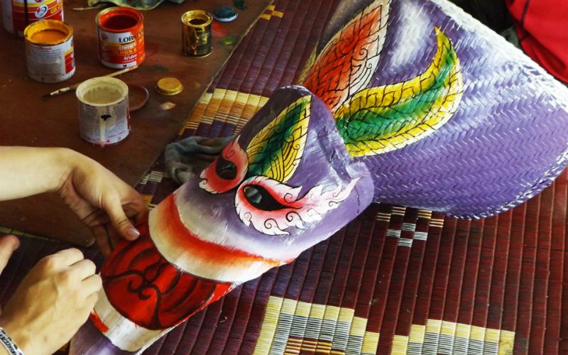 Close-up of a Phi Ta Khon mask being handcrafted in Thailand, showing a colorful painted ghost mask made from woven material with red, purple, yellow, and green details, while hands work on the mask beside open paint cans on a woven mat.